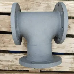 Szara, stalowa kształtka rurowa T-kształtna z kołnierzami, leżąca na drewnianej palecie. Widoczne spawy.