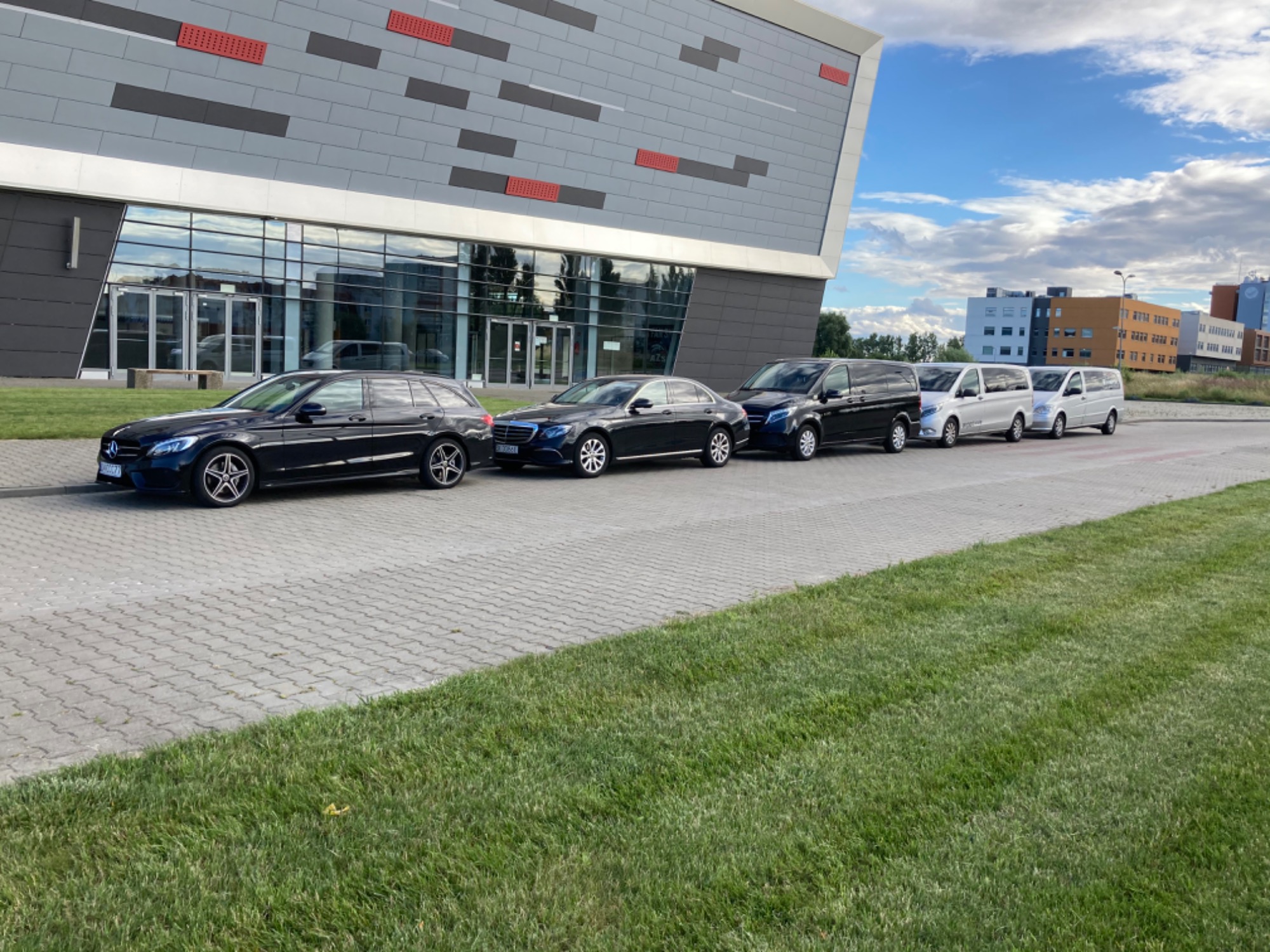 Rząd czarnych i srebrnych samochodów osobowych i vanów zaparkowanych przed nowoczesnym budynkiem z szarymi panelami i czerwonymi akcentami.
