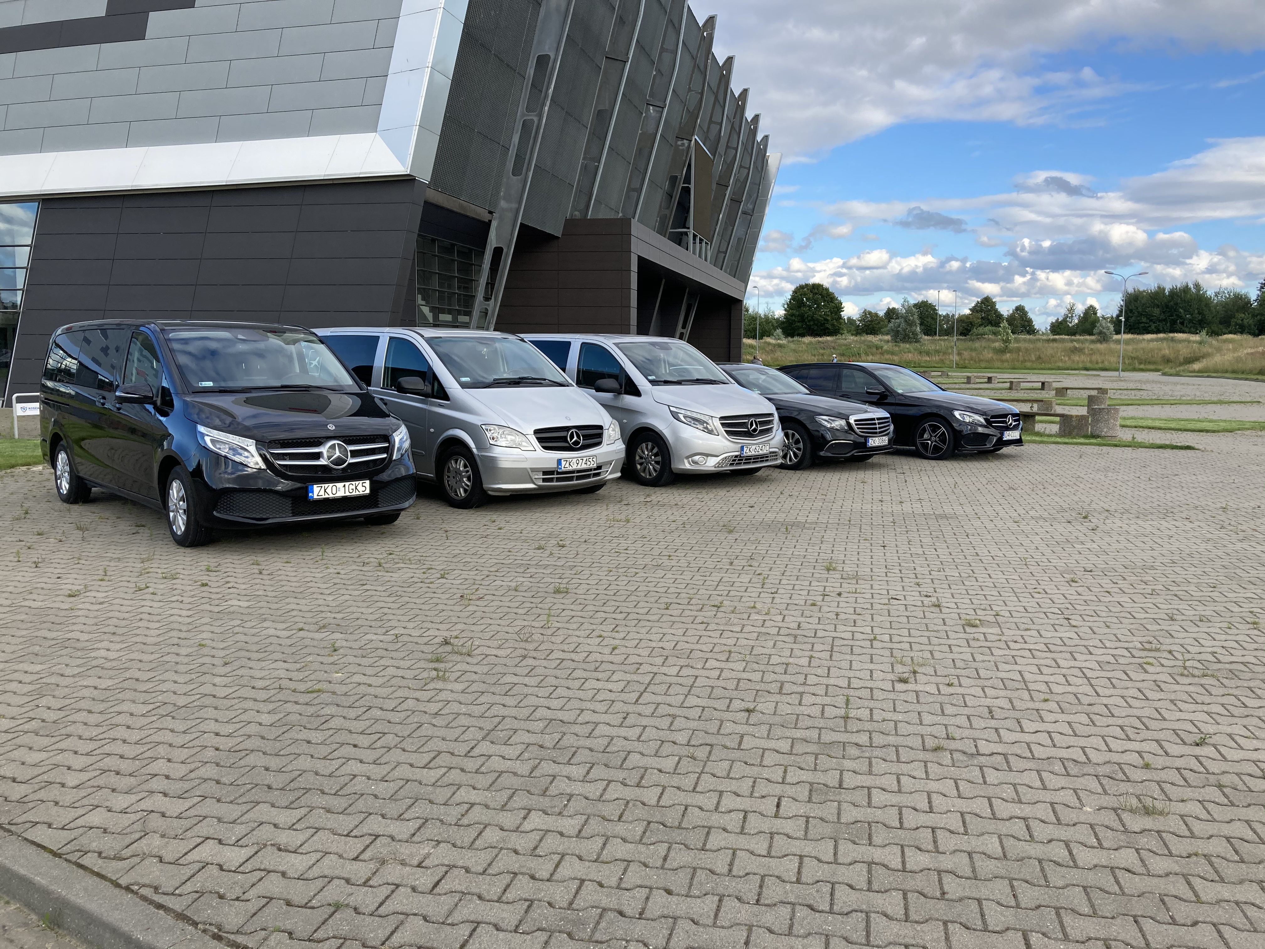 Flota samochodów Mercedes-Benz, głównie vany, zaparkowana przed nowoczesnym budynkiem z metalową elewacją w pochmurny dzień.