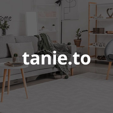 Jasne, przestronne wnętrze salonu w stylu skandynawskim z szarą sofą, białym stolikiem kawowym i drewnianym regałem. Na ścianie minimalistyczna grafika. Na pierwszym planie delikatny wzór na dywanie.