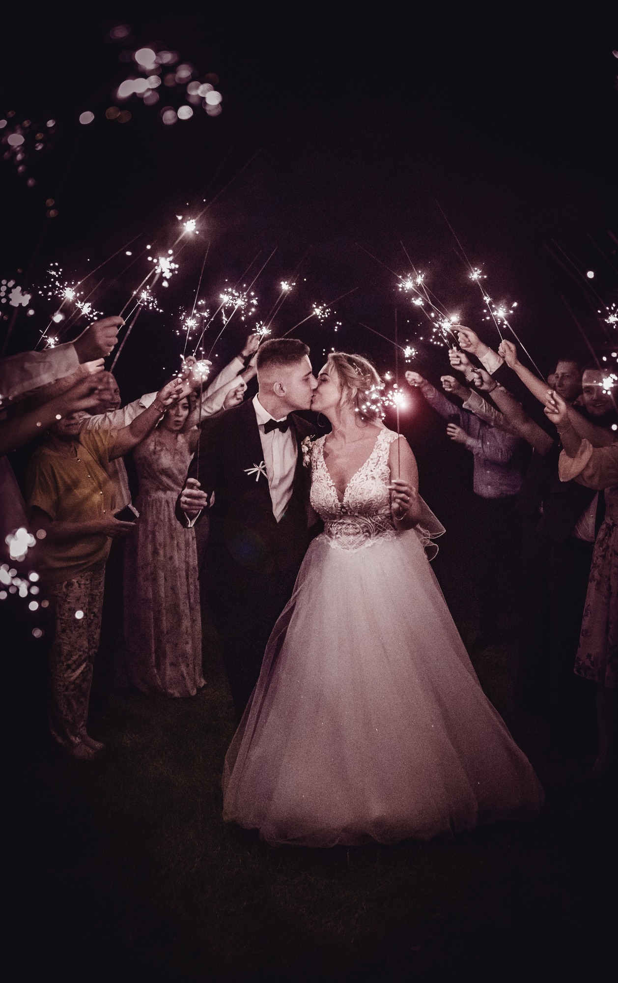 Pocałunek nowożeńców otoczonych gośćmi z zimnymi ogniami w ciemności.