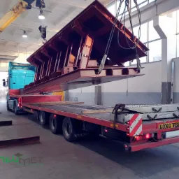 Ciężka, pomalowana na czerwono konstrukcja stalowa podnoszona przez suwnicę w hali, przygotowywana do transportu na niskopodwoziowej naczepie. Widoczny niebieski ciągnik siodłowy Volvo i żółta...