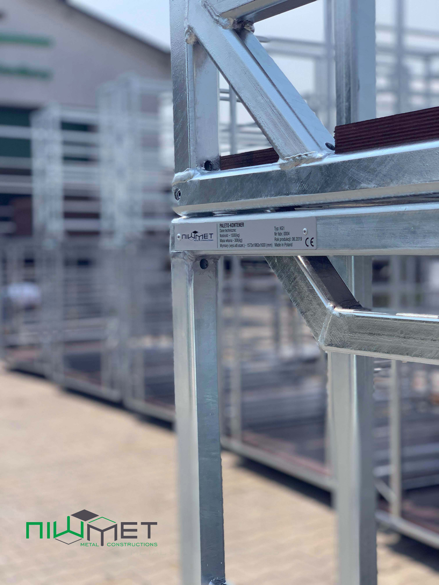 Szczegółowe ujęcie metalowej ramy kontenera paletowego, widoczne spawy i tabliczka znamionowa z danymi technicznymi, w tle inne kontenery.