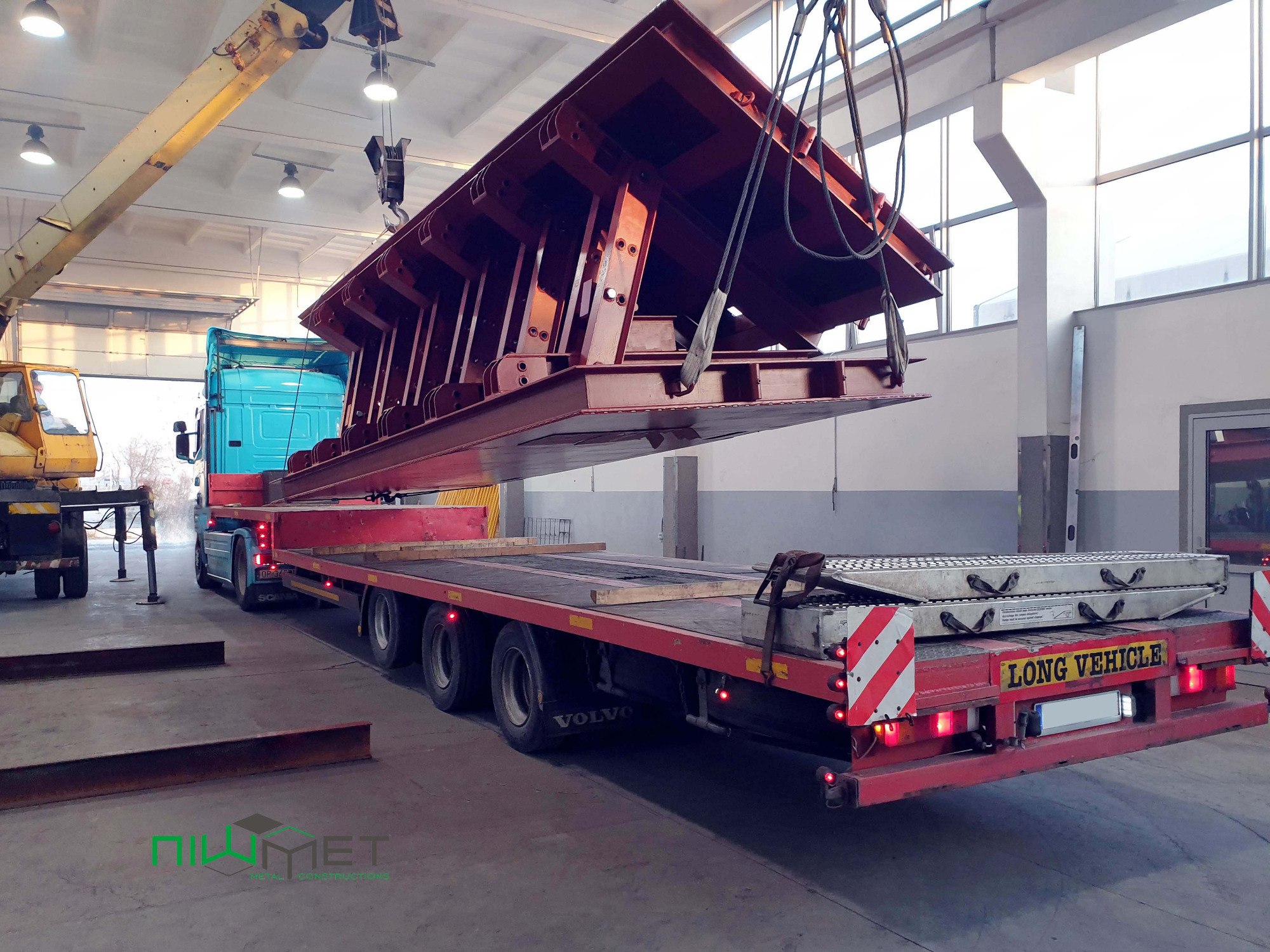 Ciężka, pomalowana na czerwono konstrukcja stalowa podnoszona przez suwnicę w hali, przygotowywana do transportu na niskopodwoziowej naczepie. Widoczny niebieski ciągnik siodłowy Volvo i żółta...