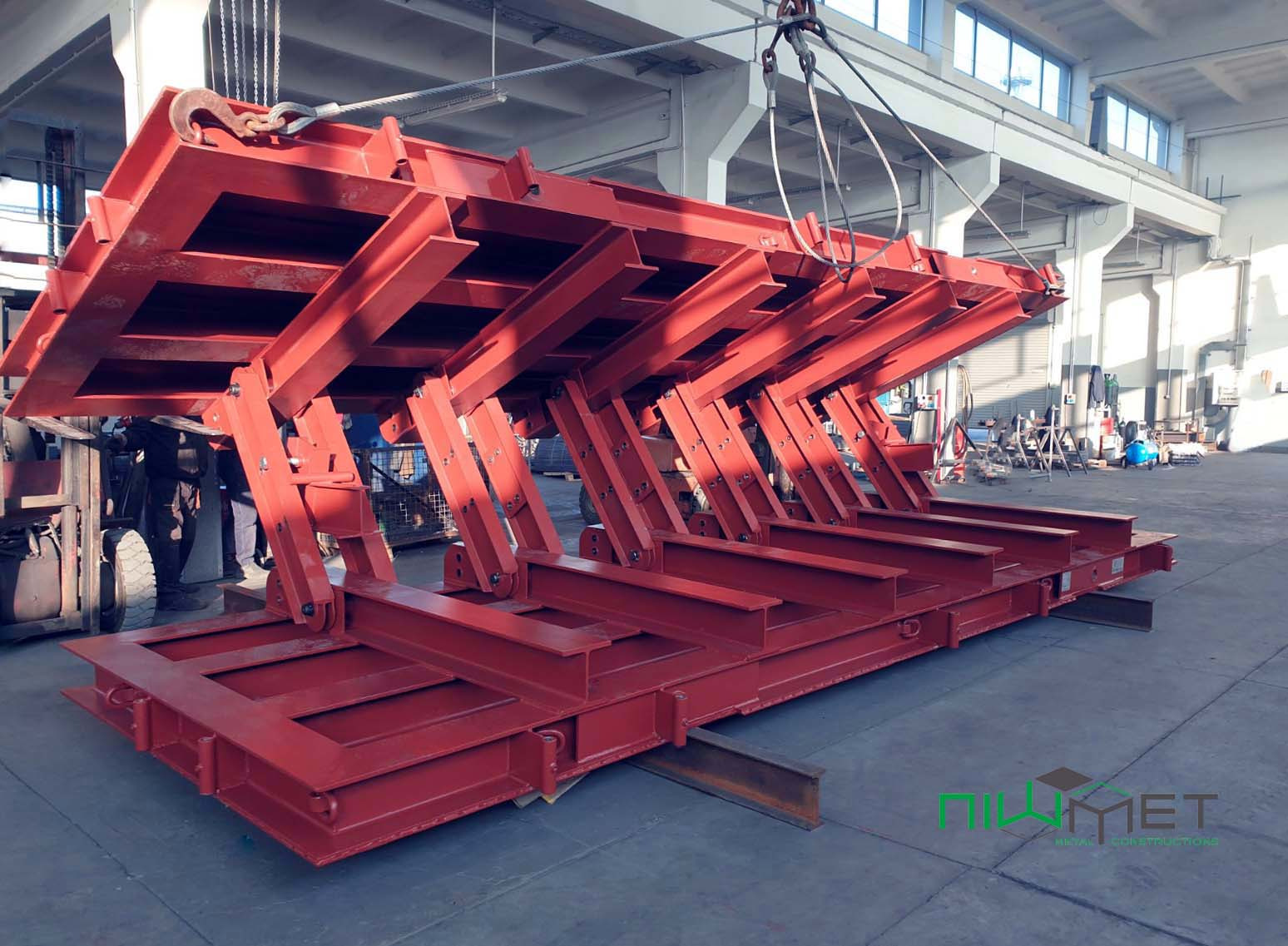 Czerwona, stalowa konstrukcja podnośnika nożycowego w hali produkcyjnej, podwieszona na linach z hakiem, widoczny wózek widłowy i logo firmy.