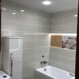 Wykończona łazienka z szarymi płytkami, podświetlanym panelem dekoracyjnym nad wanną, białą zabudową i pralką.