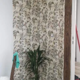 Ściana pokryta wzorzystą tapetą z motywami tropikalnymi, z widoczną drewnianą belką, doniczką z rośliną i akcesoriami malarskimi, sugerującymi prace wykończeniowe.