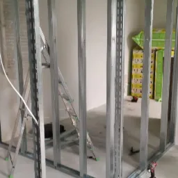 Metalowy szkielet ścianki działowej z profili stalowych, widoczna drabina aluminiowa i wkrętarka akumulatorowa, w tle płyty gipsowo-kartonowe.