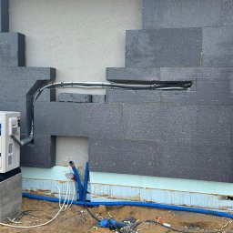 ECOTERM CO SP. Z O.O. - Biała pompa ciepła Haier na betonowych podstawach, z żółtą rurą i niebieskimi przewodami, zamontowana przy ocieplonej ścianie budynku z otwartymi drzwiami do wnętrza.