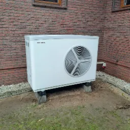 Zewnętrzna jednostka pompy ciepła marki Stiebel Eltron, umieszczona na betonowych podstawach przy ceglanej ścianie domu, widoczny kabel zasilający i otoczenie z kamieniami i ziemią.