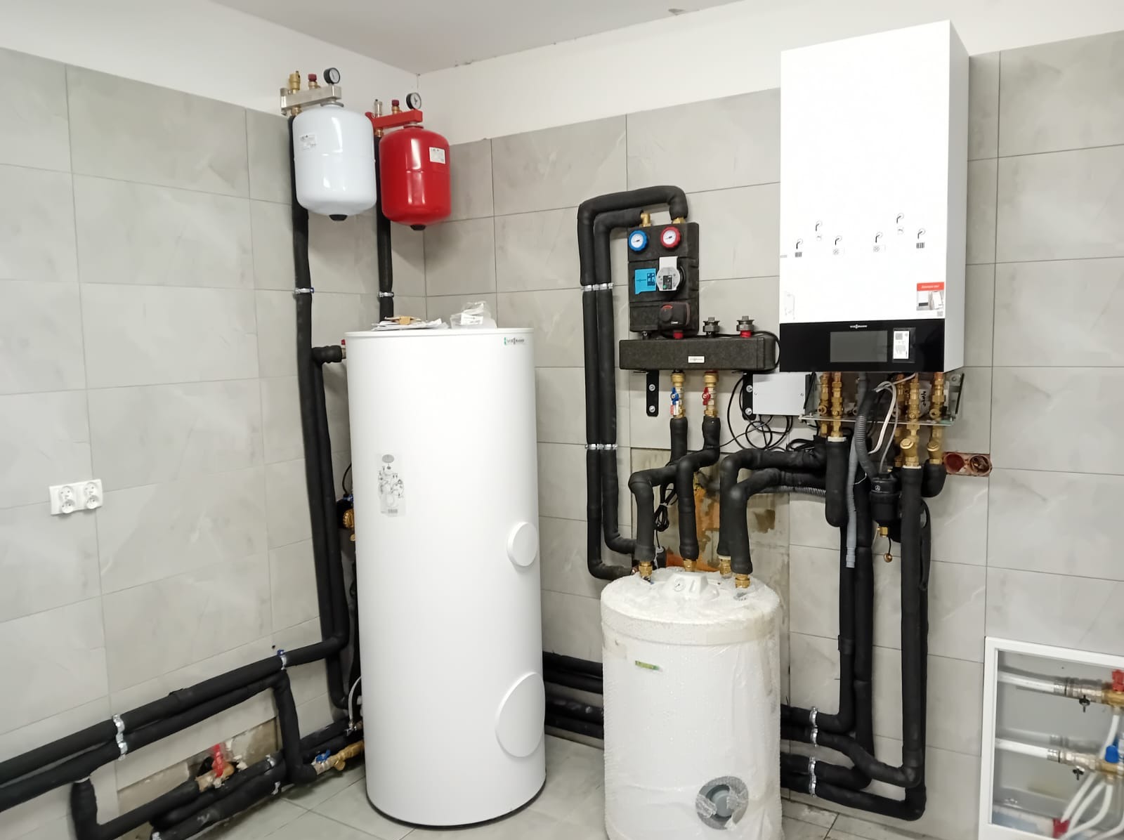 Złożona instalacja grzewcza z kotłem, zbiornikami i rurami w pomieszczeniu z szarymi płytkami. Widoczne elementy instalacji CO i CWU.