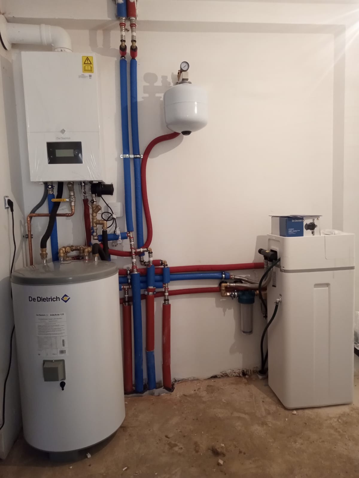 Biały piec gazowy De Dietrich, zbiornik na wodę i zmiękczacz wody połączone rurami w kolorze niebieskim i czerwonym na tle białej ściany i surowej podłogi.