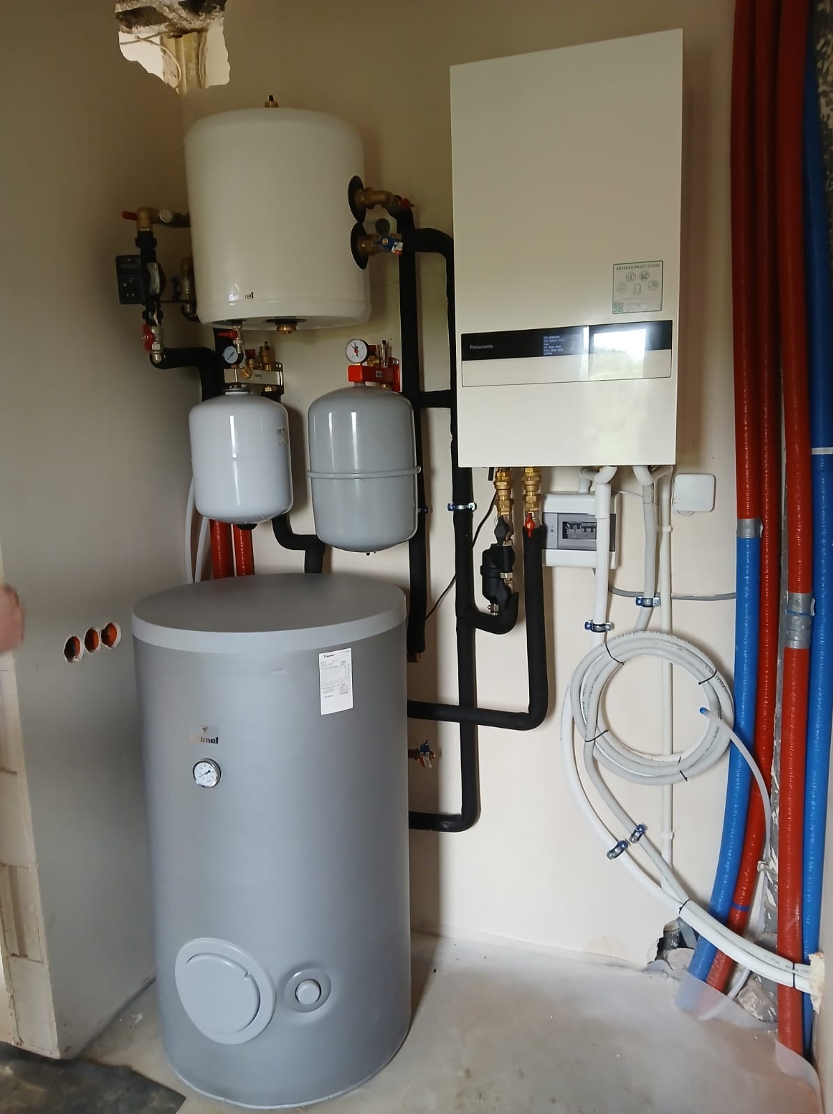 Zestaw instalacji grzewczej z kotłem gazowym, zbiornikiem i rurami w kolorach niebieskim i czerwonym. Widoczne elementy instalacji i czarne rury. Świeżo po montażu.