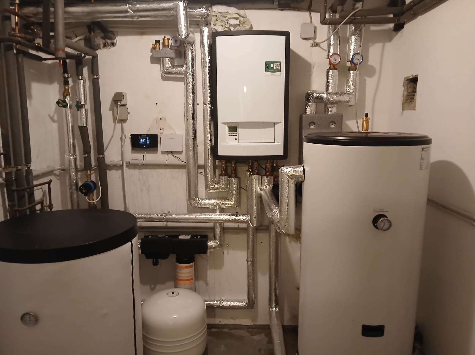 Biały piec gazowy z czarną obudową, zbiorniki wody i rury w kotłowni. Widoczny panel sterowania i filtry. Instalacja po montażu, rury zaizolowane.