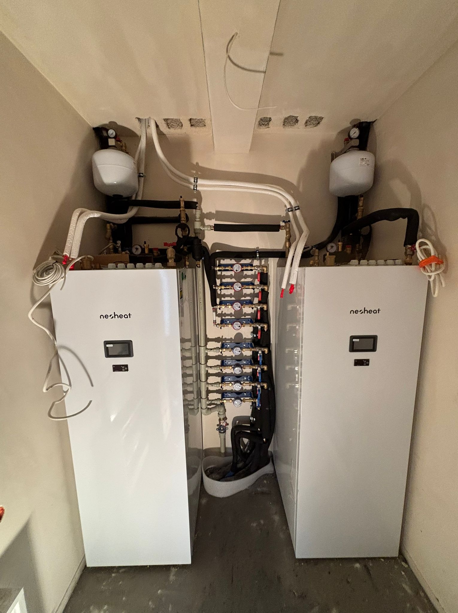 Dwie białe pompy ciepła Nesheat z podłączonymi rurami w pomieszczeniu. Widoczne zawory, manometry i zbiorniki wyrównawcze. Instalacja grzewcza w kotłowni.