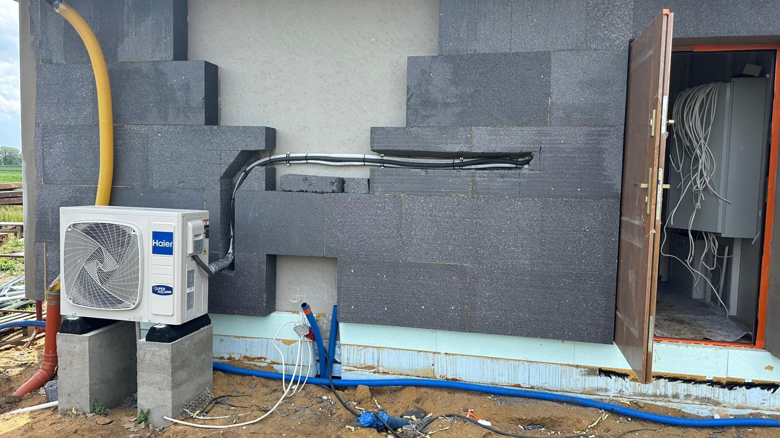 Biała pompa ciepła Haier na betonowych podstawach, z żółtą rurą i niebieskimi przewodami, zamontowana przy ocieplonej ścianie budynku z otwartymi drzwiami do wnętrza.