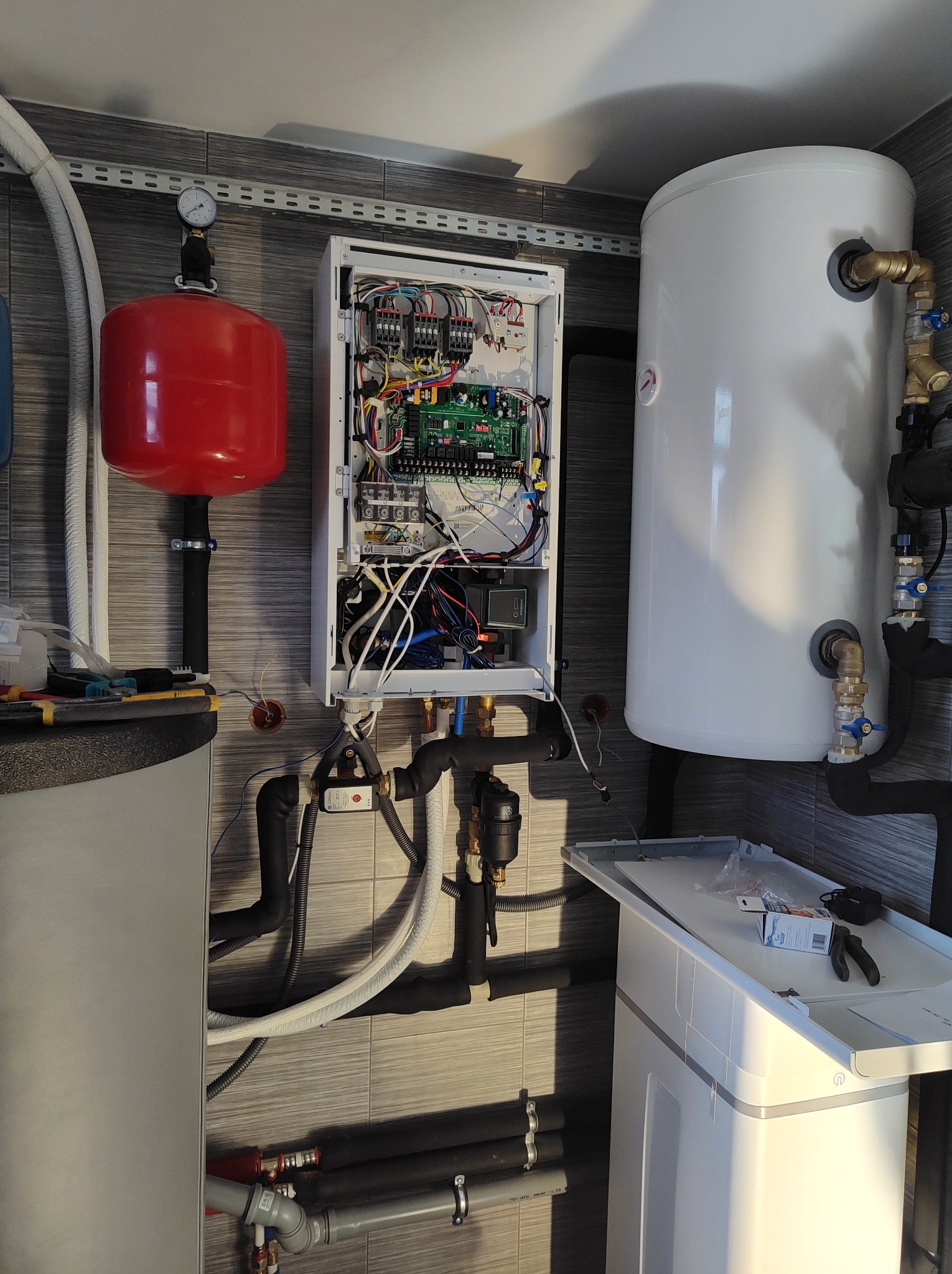 Złożona instalacja grzewcza z kotłem, zbiornikiem i czerwonym naczyniem wzbiorczym na tle szarej ściany z płytek. Widoczne rury, kable i narzędzia.