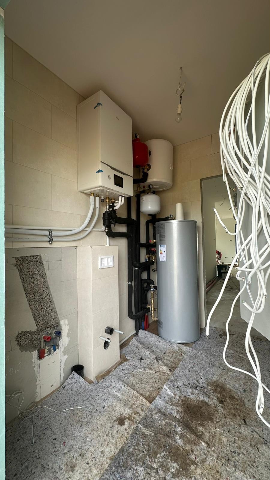 Biały piec gazowy kondensacyjny z czarnymi rurami, zbiornik wody i instalacja w pomieszczeniu wyłożonym płytkami. Podłoga zabezpieczona materiałem ochronnym.