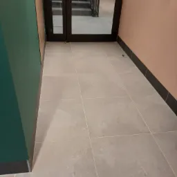 Jasnoszare płytki podłogowe w korytarzu z zieloną ścianą po lewej i różową po prawej, widoczne schody przez szklane drzwi na wprost.