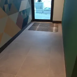 Wnętrze korytarza z beżowymi płytkami podłogowymi, metalową wycieraczką przy wejściu i ścianą z geometrycznym wzorem w odcieniach niebieskiego, beżu i fioletu.