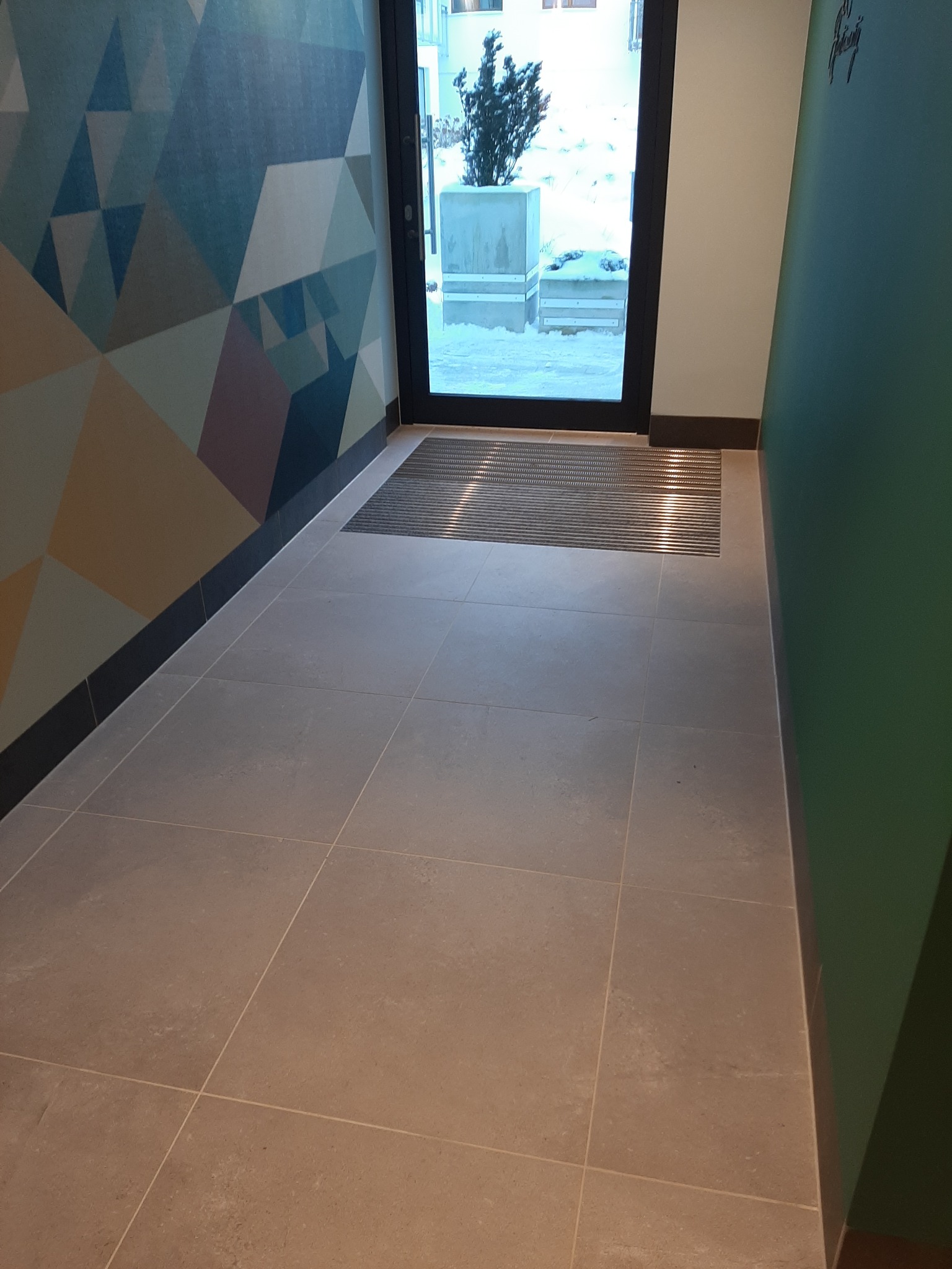 Wnętrze korytarza z beżowymi płytkami podłogowymi, metalową wycieraczką przy wejściu i ścianą z geometrycznym wzorem w odcieniach niebieskiego, beżu i fioletu.