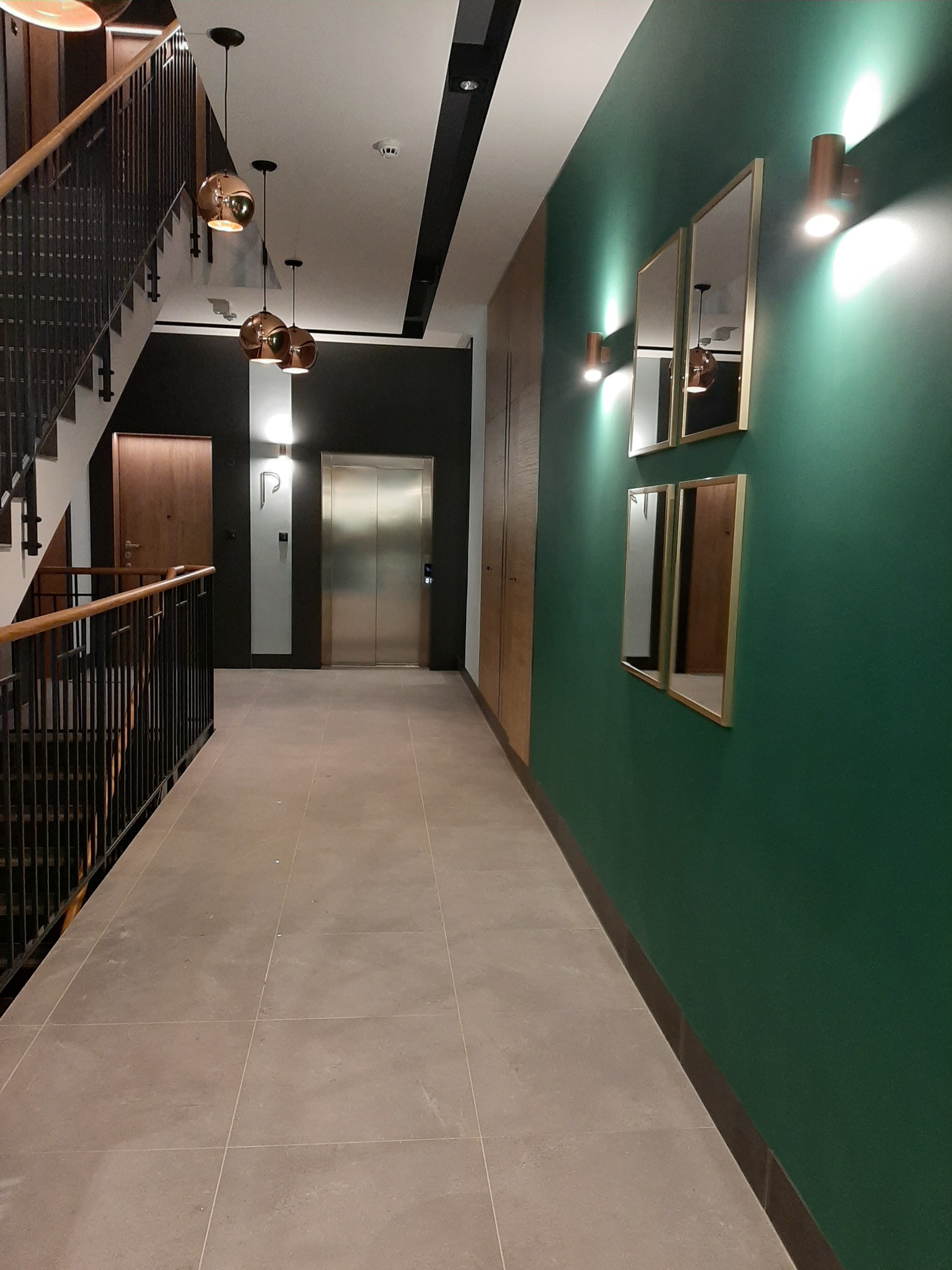 Korytarz w nowoczesnym budynku z zieloną ścianą, lustrami w złotych ramach i schodami z czarną balustradą. Na suficie wiszą miedziane lampy, a w tle widoczna jest winda.