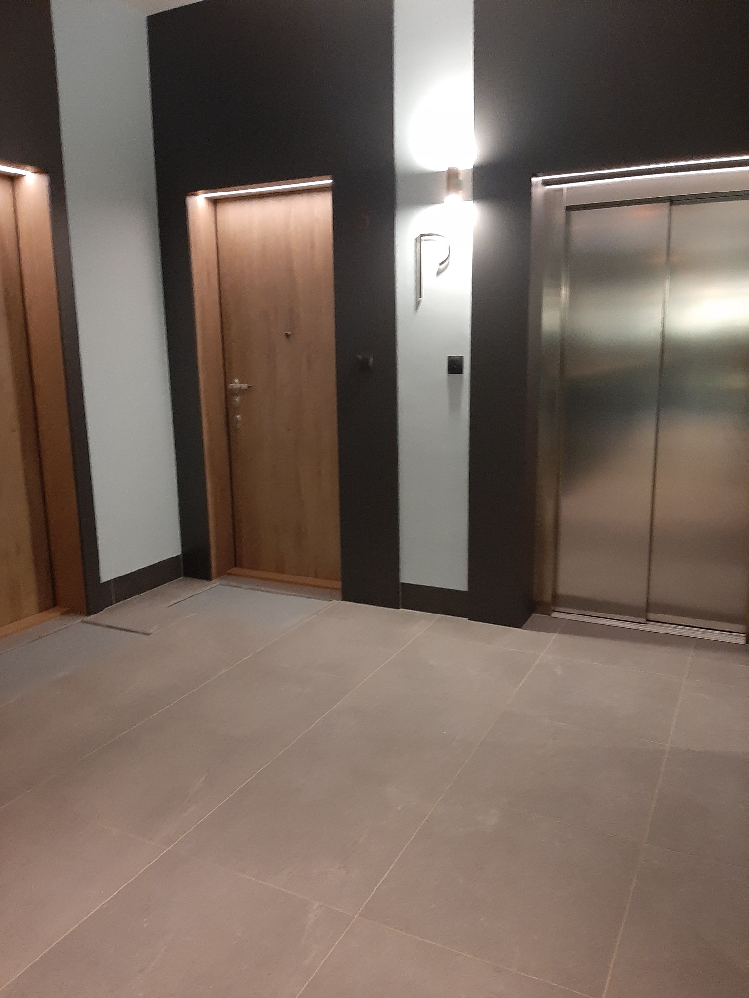 Korytarz w budynku z drzwiami wejściowymi do mieszkań i windą, widoczne płytki podłogowe w odcieniach beżu.