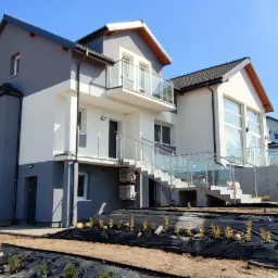 Nowoczesny dom dwukondygnacyjny z balkonem i szklanymi balustradami, świeżo po zakończeniu prac elewacyjnych, z zagospodarowanym terenem wokół, w słoneczny dzień.