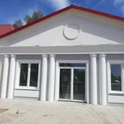 Nowo wykończona elewacja budynku z kolumnami, okna zabezpieczone folią, czerwony dach, widoczne niedociągnięcia na podłożu.