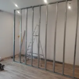 Wnętrze w trakcie montażu ścianki działowej z metalowych profili, drabina oparta o konstrukcję, narzędzia i akcesoria na podłodze, widoczne okno z żaluzjami.