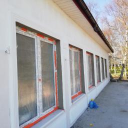 NO I FAJNIE DANIEL WANAGO - Świeżo otynkowana elewacja budynku z oknami zabezpieczonymi folią, widoczne detale architektoniczne i elementy wykończeniowe, na pierwszym planie niebieski worek i fragment żółtej gąbki.