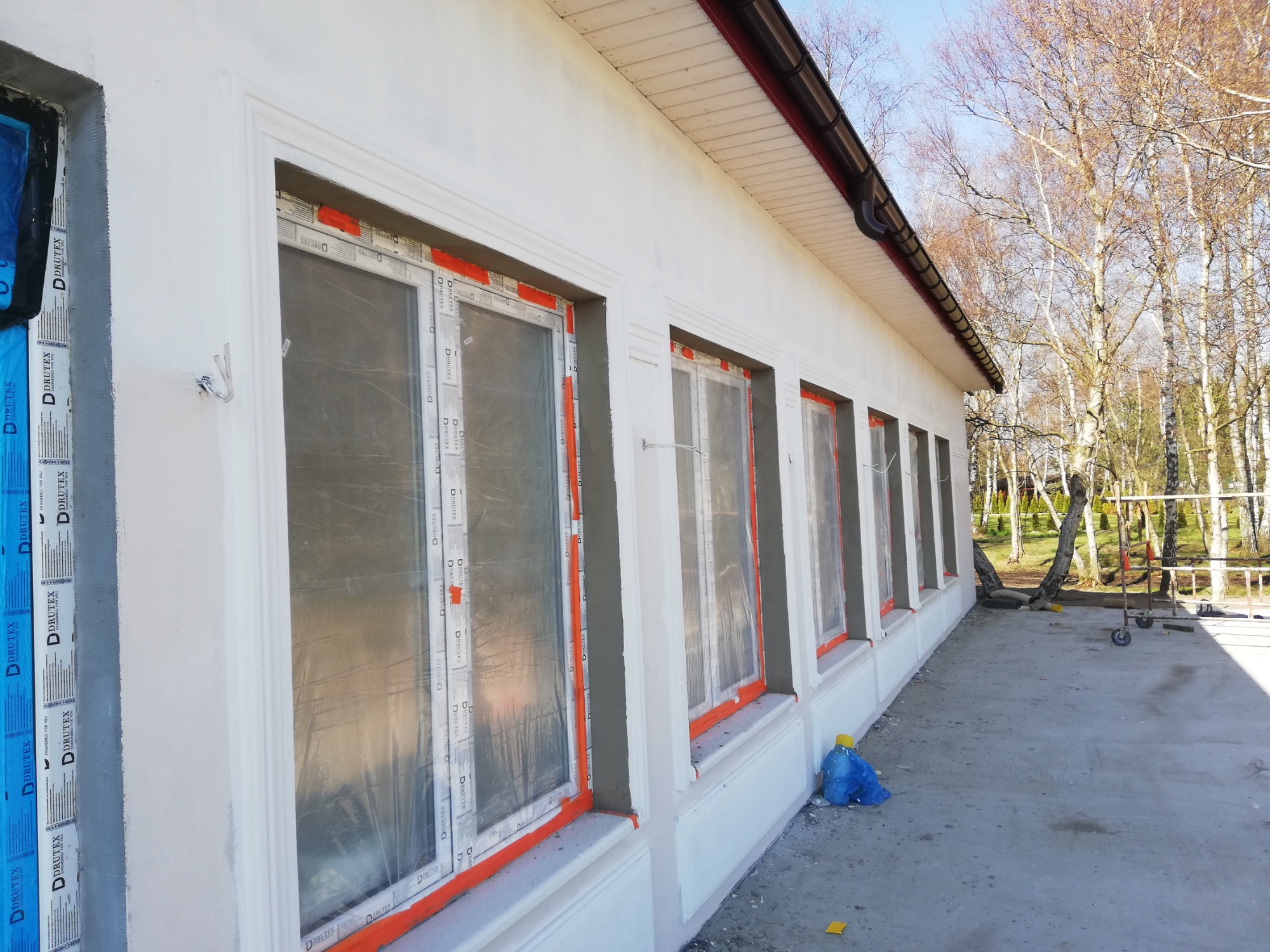 Świeżo otynkowana elewacja budynku z oknami zabezpieczonymi folią, widoczne detale architektoniczne i elementy wykończeniowe, na pierwszym planie niebieski worek i fragment żółtej gąbki.