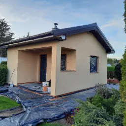 Niski budynek po tynkowaniu elewacji, zabezpieczony folią malarską, widoczne puszki z farbą i narzędzia.