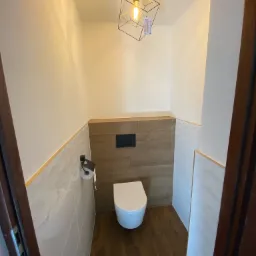 Mała toaleta z drewnianą podłogą i częściowo drewnianą ścianą, nad którą znajduje się spłuczka, a na ścianie po lewej stronie wisi podajnik na papier toaletowy. Ściany wyłożone są białymi płytkami...