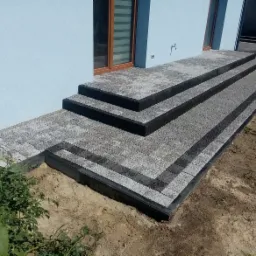 Schody wejściowe do domu wyłożone szarą kostką brukową z czarnymi obrzeżami, widok z boku na tle jasnoniebieskiej elewacji budynku.