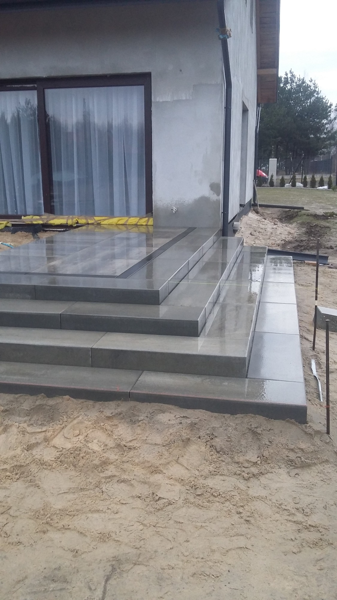 Nowe, szare schody wejściowe z płytek, prowadzące do surowego domu, w tle okno z zasłoniętymi zasłonami i piasek.