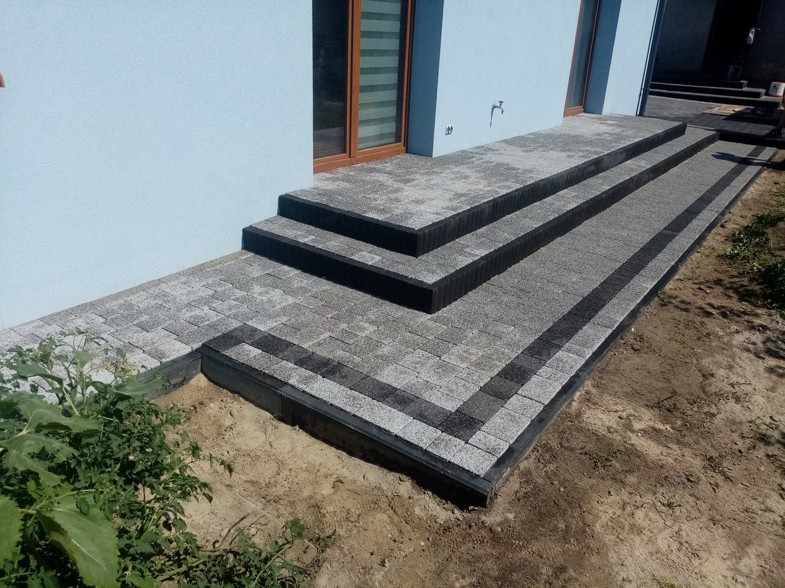 Schody wejściowe do domu wyłożone szarą kostką brukową z czarnymi obrzeżami, widok z boku na tle jasnoniebieskiej elewacji budynku.