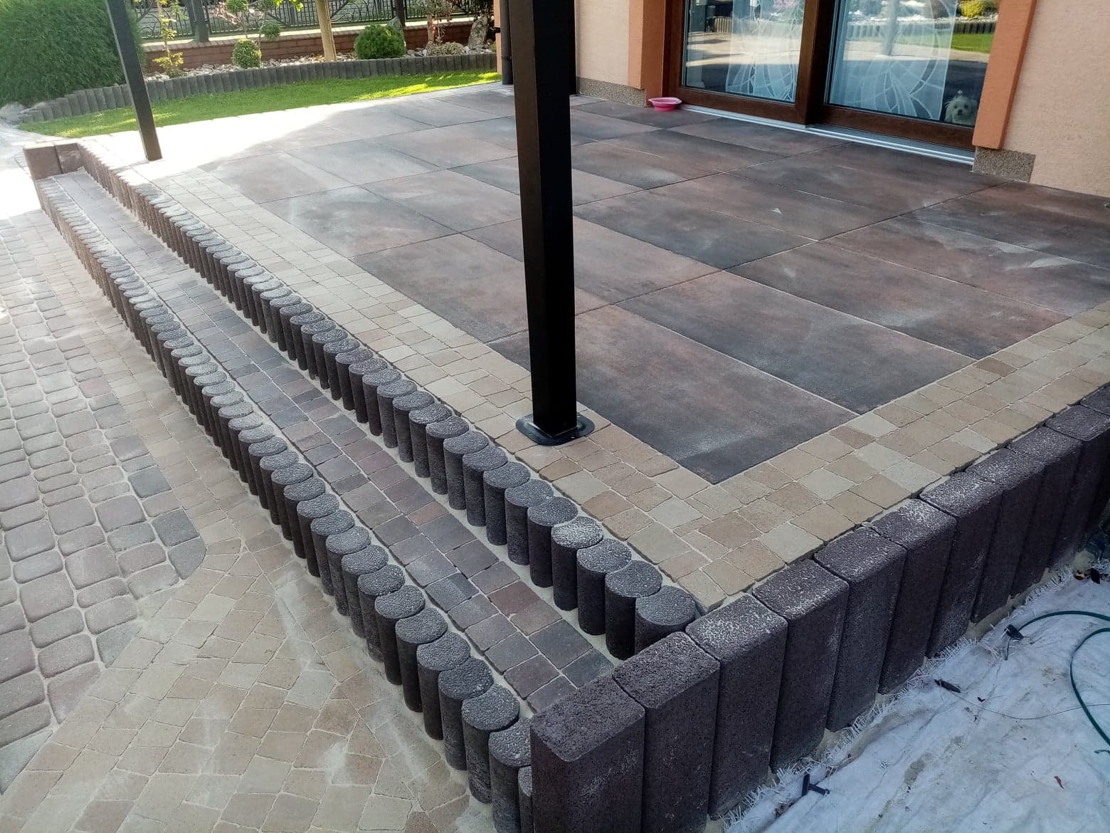 Nowoczesny taras z kostki brukowej w odcieniach brązu i beżu, z niskimi schodami obłożonymi dekoracyjną kostką cylindryczną, widok na dom z przesuwnymi drzwiami.