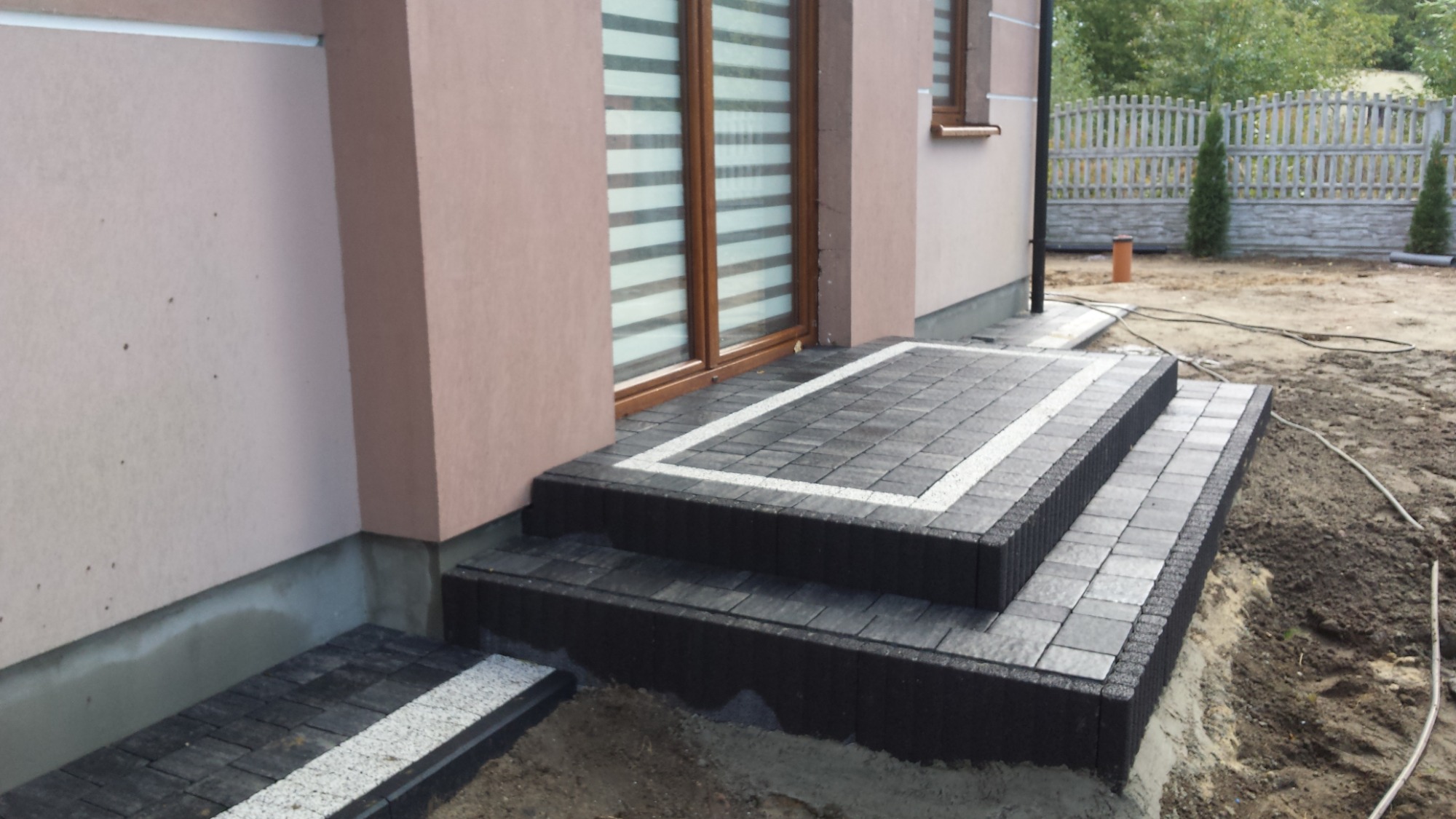 Schody wejściowe do domu wyłożone ciemną kostką brukową z jasnymi obramowaniami, widoczne okno z roletami i fragment ogrodu z ogrodzeniem.
