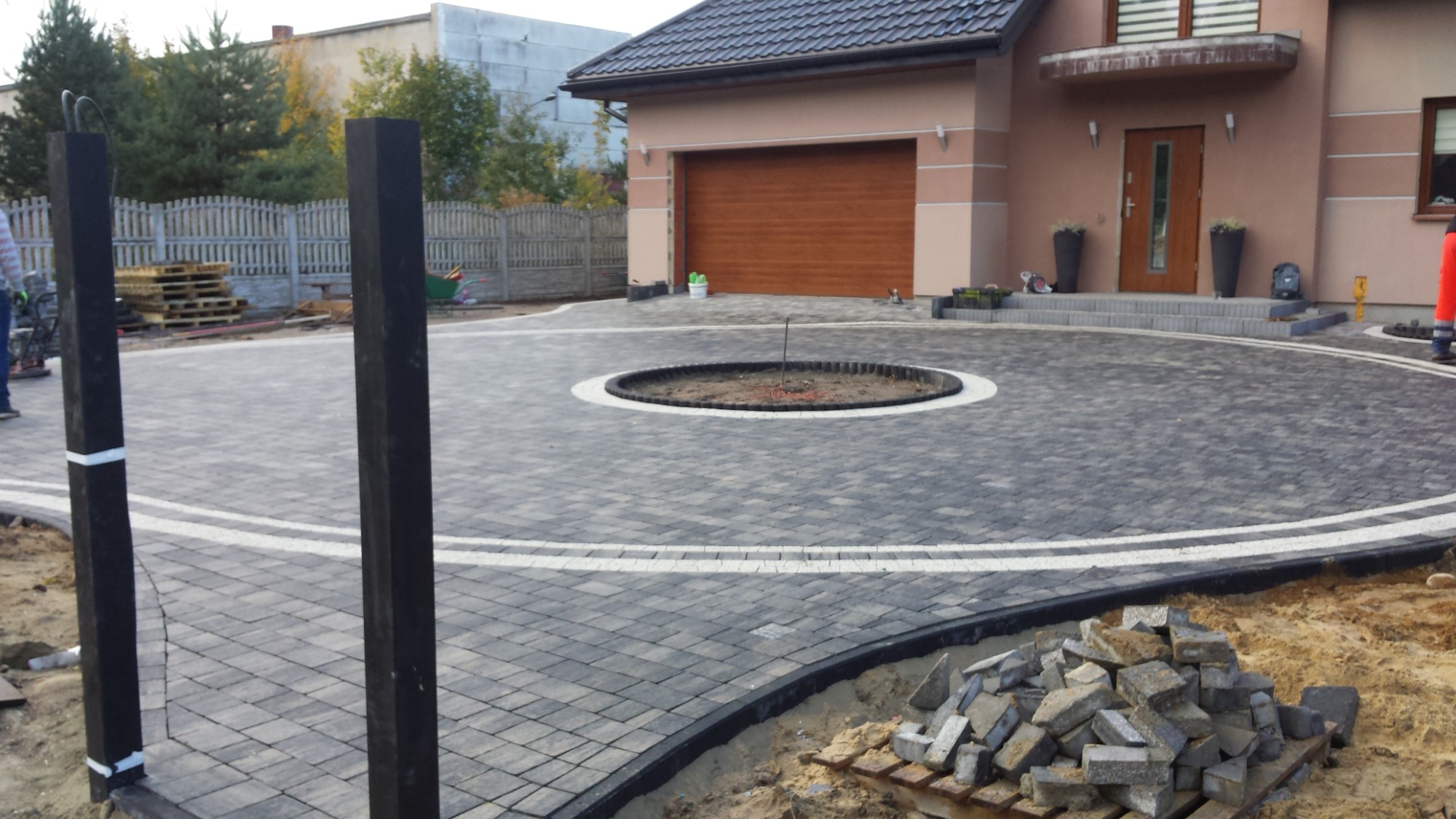 Okazały podjazd z szarej kostki brukowej ułożonej w okrągły wzór przed domem z garażem, z widocznym przygotowanym miejscem na trawnik pośrodku i stertą kostki do wykończenia.