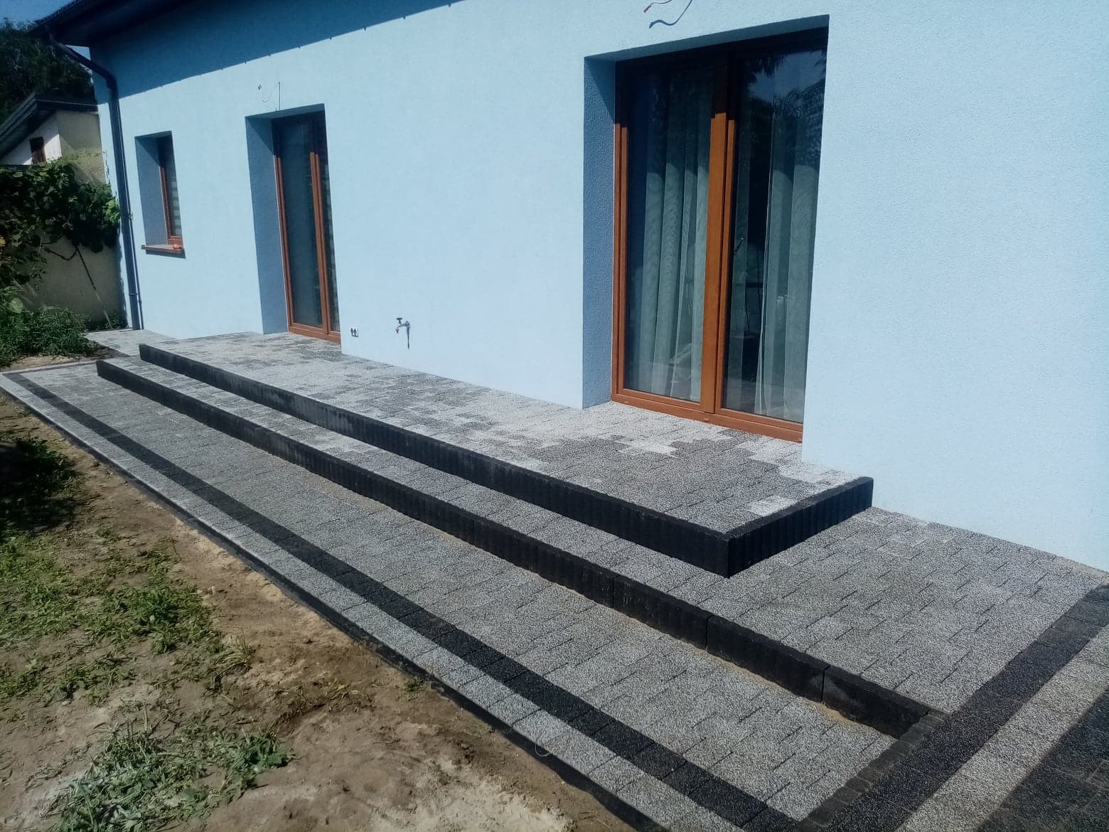 Nowe schody i chodnik z szarej kostki brukowej prowadzące do domu z błękitną elewacją i brązowymi oknami.