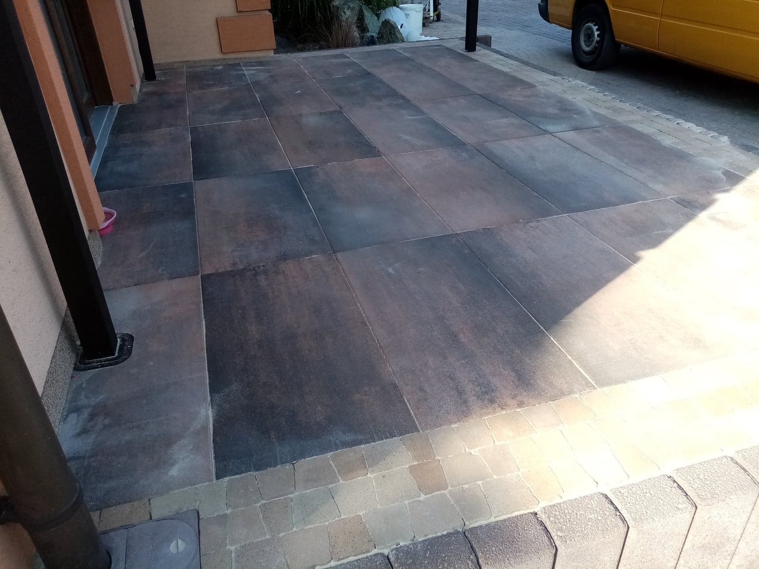 Ułożona, duża, ciemna kostka brukowa na podjeździe, obrzeża z jaśniejszych, mniejszych kostek, widoczny fragment żółtego samochodu.