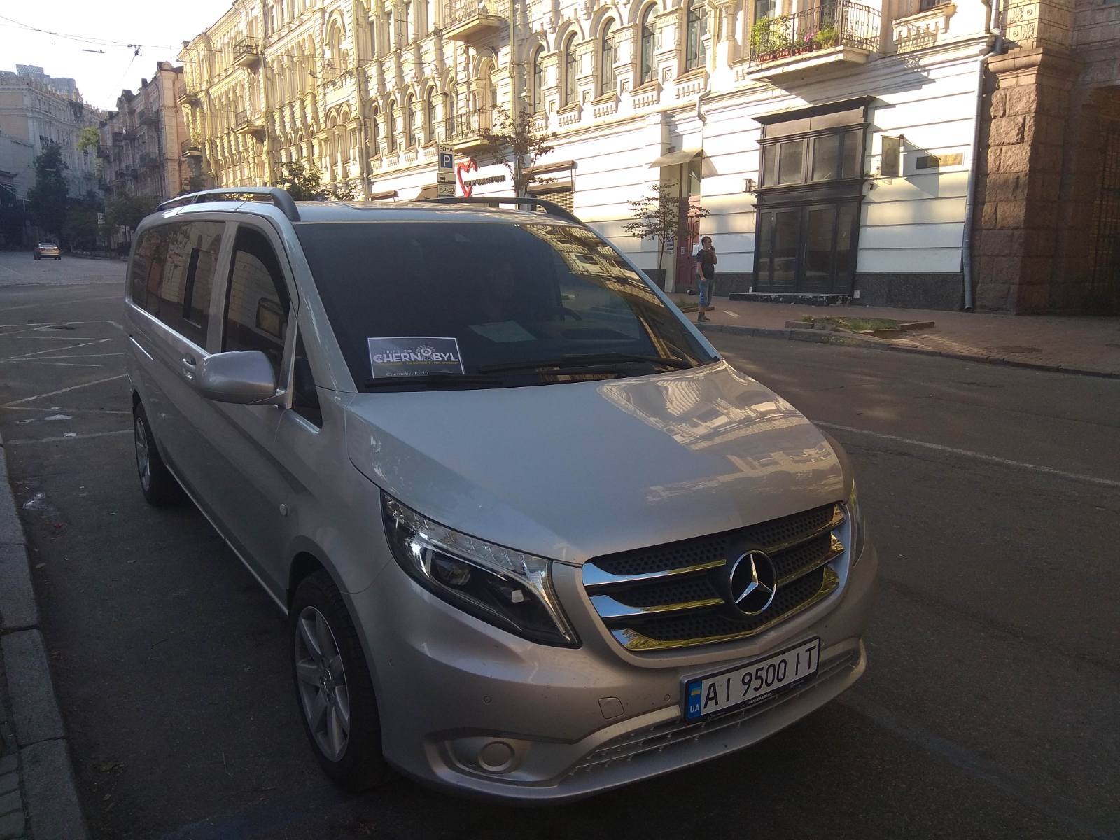 Srebrny Mercedes-Benz Vito z naklejką 'Chernobyl' na przedniej szybie, zaparkowany na ulicy z klasyczną architekturą budynków w tle.