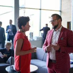 Kobieta w czerwonej sukience rozmawia z mężczyzną w bordowym garniturze na tle biurowego wnętrza z sofami i dużymi oknami.