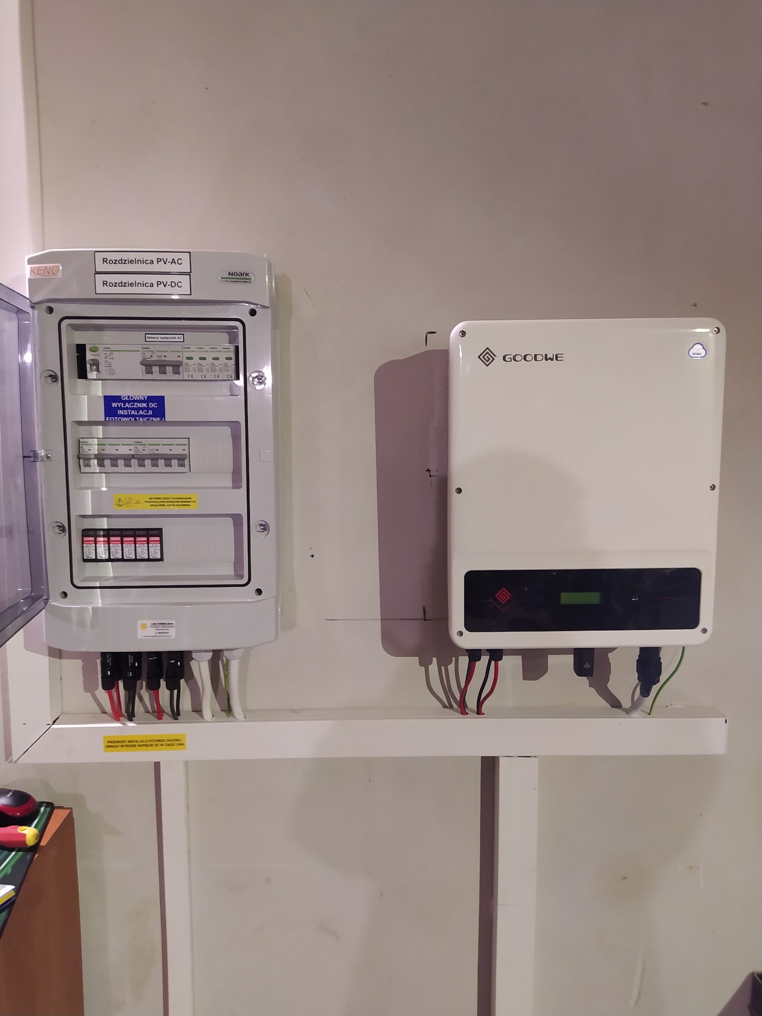 Rozdzielnica PV-AC i PV-DC firmy KENO z otwartymi drzwiczkami, obok biały inwerter fotowoltaiczny GoodWe na białej półce przy ścianie.