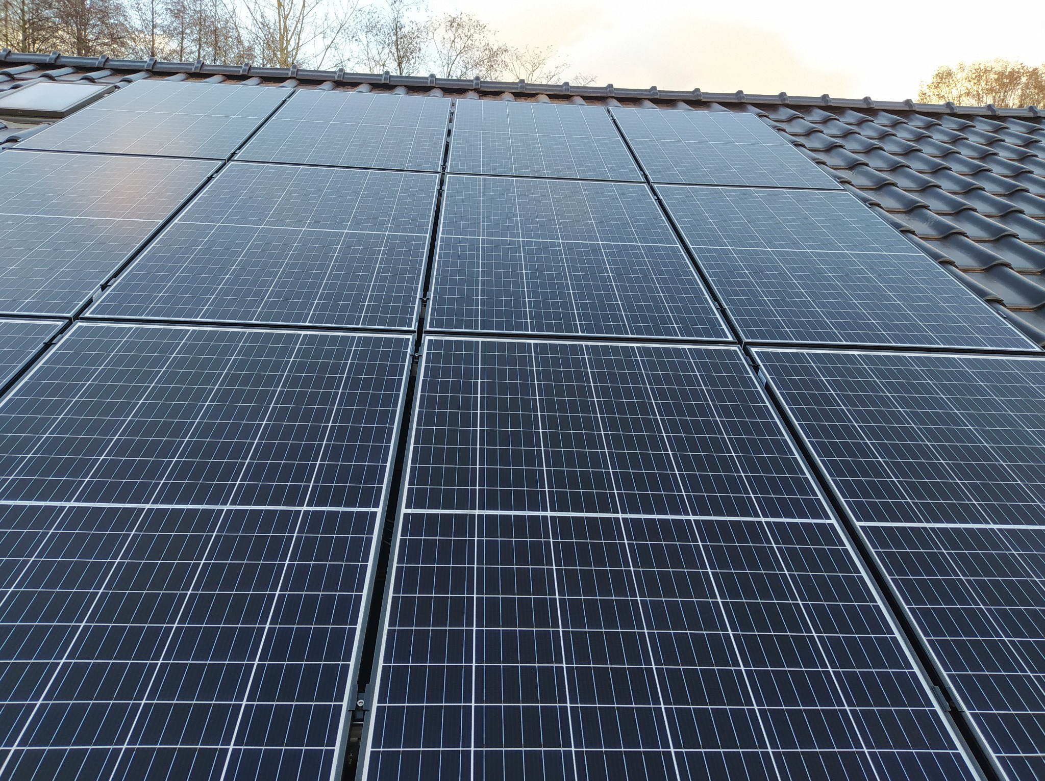 Panele fotowoltaiczne zamontowane na dachu pokrytym ciemną dachówką, widoczne okno dachowe i drzewa w tle.