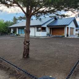 Piotr - Przygotowany teren pod trawnik z systemem nawadniania, widok na dom z garażem i motocyklem. W tle zieleń drzew i krzewów, częściowo widoczne ogrodzenie.