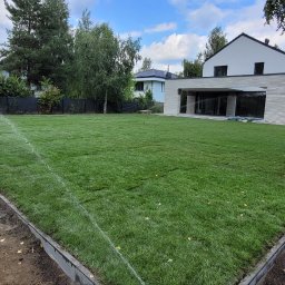 Piotr - Nawadnianie trawnika z rolki w ogrodzie nowoczesnego domu. Widoczne zraszacze i świeżo ułożona trawa. Granica trawnika wykończona obrzeżem.