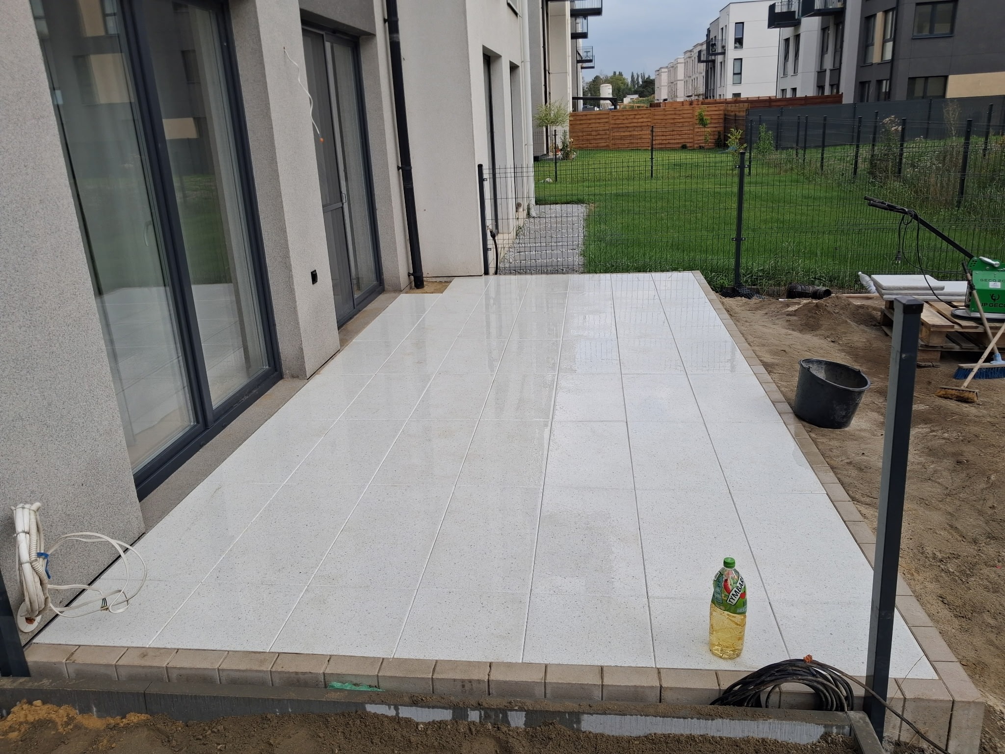 Wyłożony jasnymi płytkami taras przy nowoczesnym domu z dużymi oknami. Widoczne narzędzia i materiały budowlane, sugerujące trwające prace wykończeniowe.