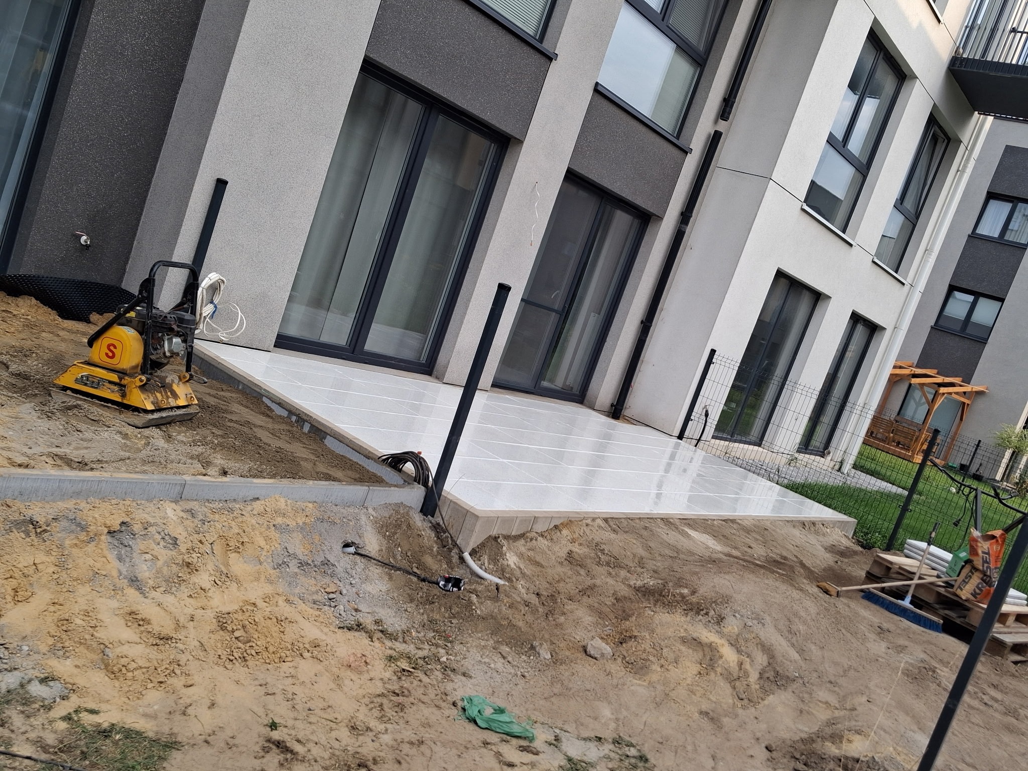 Układanie jasnych płyt tarasowych przy nowoczesnym budynku. W tle widoczna zagęszczarka i elementy konstrukcyjne pergoli. Teren wokół w trakcie prac ziemnych.